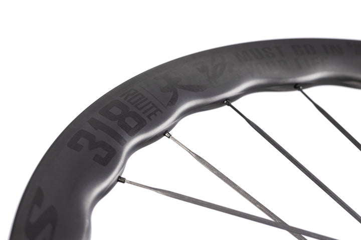 UNAAS Pro2 carbon wheel with wavy rim, dark 'ROUTE 318' branding and 'MUST GO IN YOUR LIFE' text.