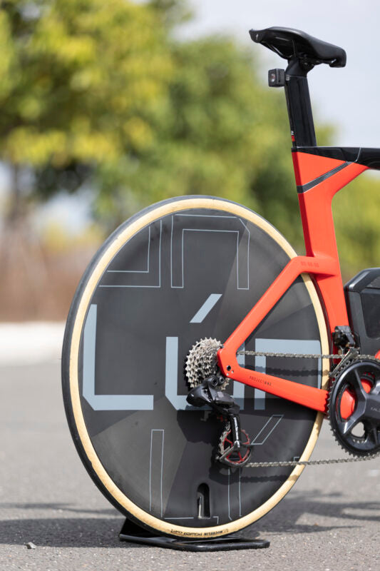 Lún Kronos disc wheel on red time trial bike, matte dark gray carbon fiber with LÚN logo. For track racing.